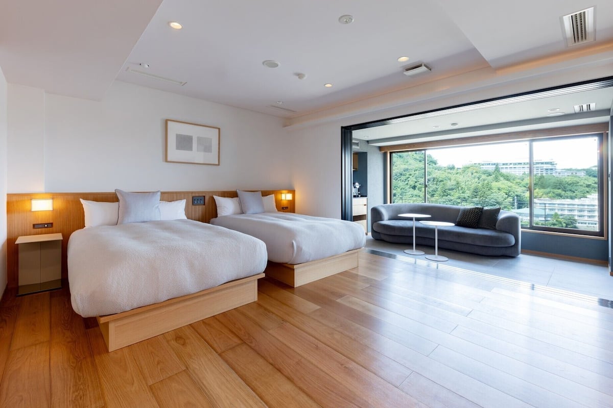 Une chambre d’hôtel moderne avec deux lits, un canapé, une petite table et une grande fenêtre donnant sur la ville.