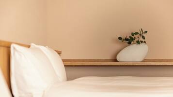 A neatly made bed with white bedding, a wooden headboard, and a white vase with green leaves on a wooden shelf against a beige wall.