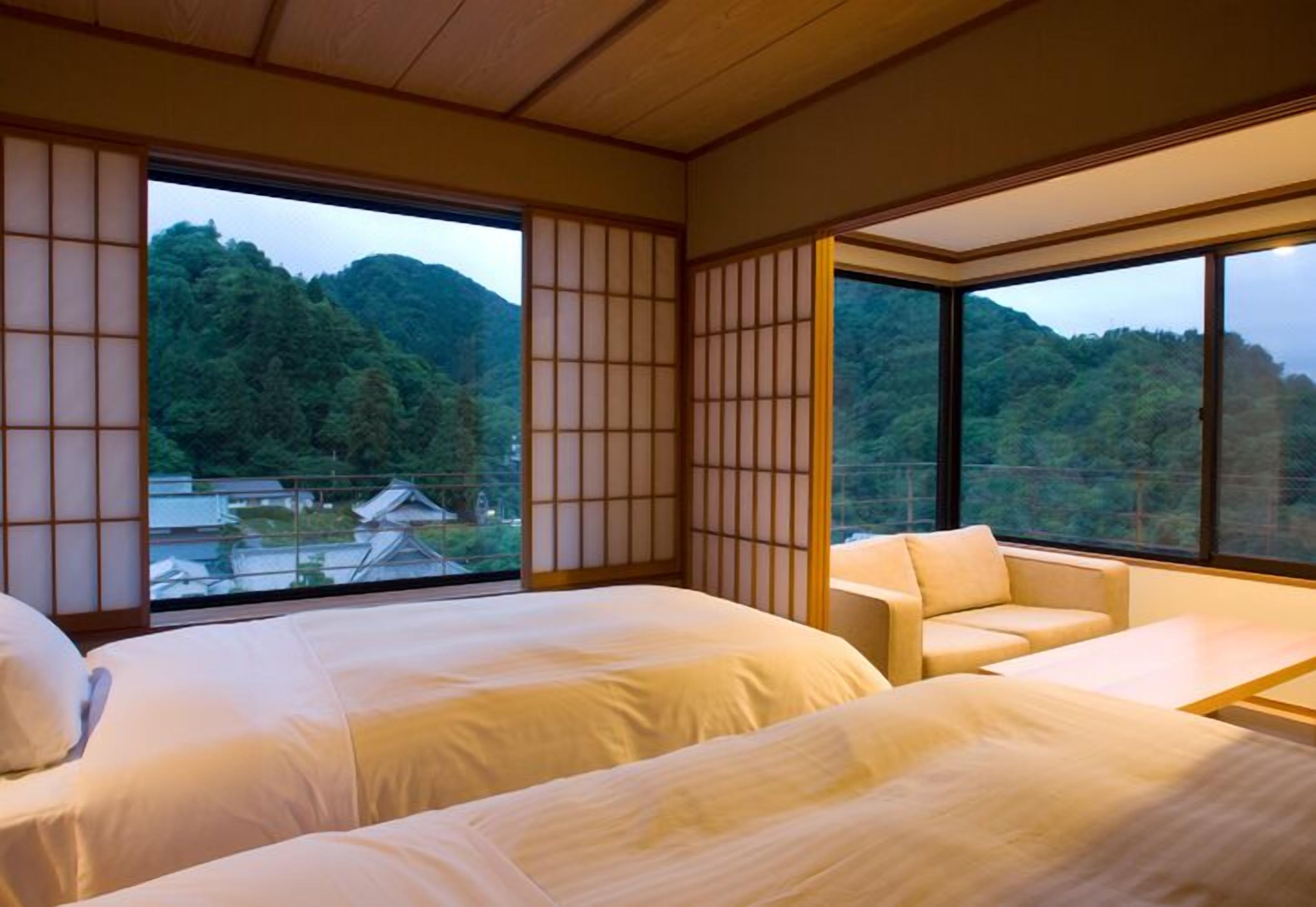 Habitación de hotel con dos camas, un sofá y grandes ventanales con vistas a una montaña boscosa.