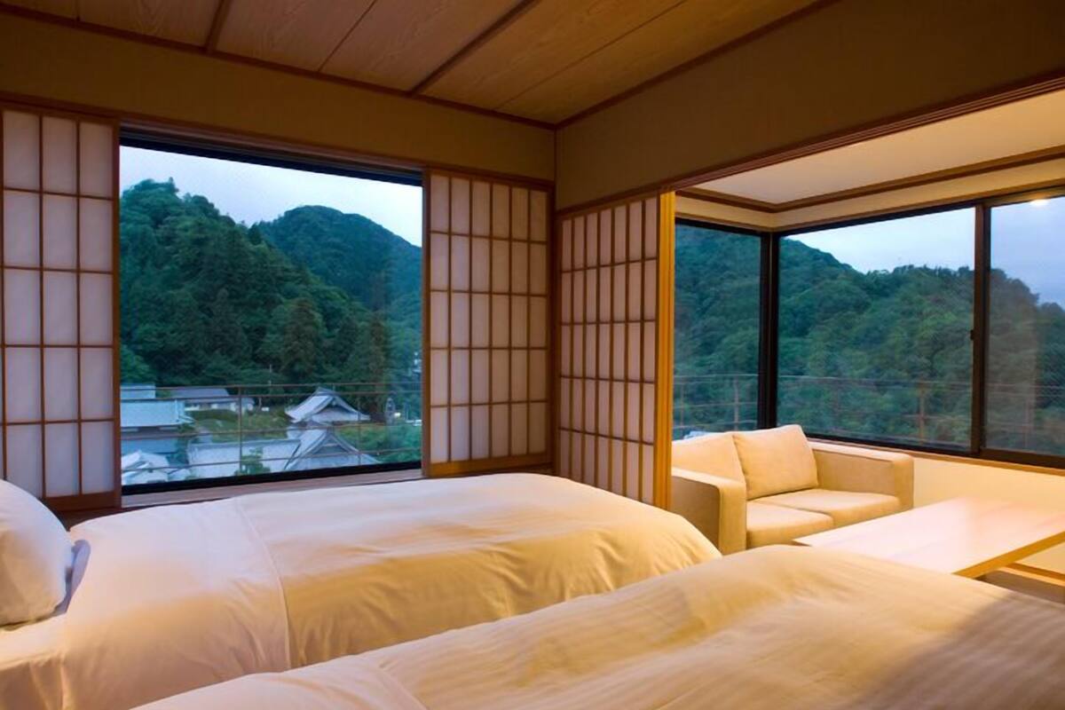 Une chambre d’hôtel avec deux lits, un canapé et de grandes fenêtres donnant sur une montagne boisée.