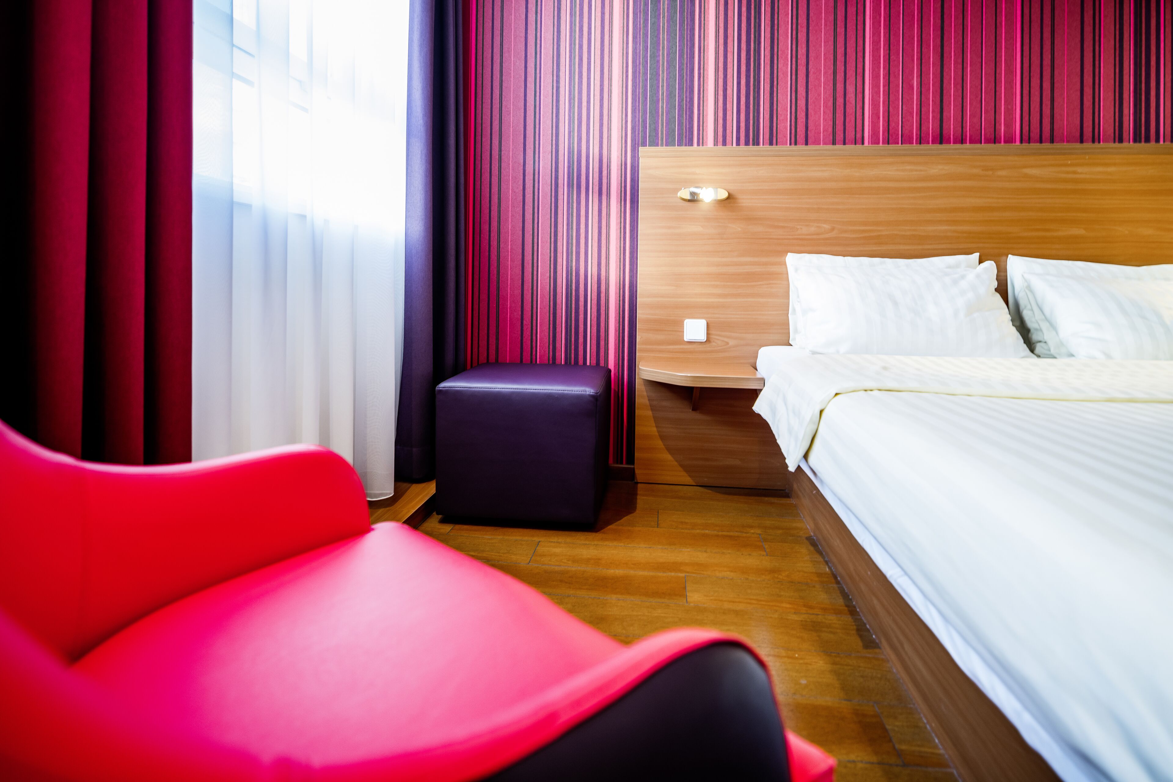 A hotel room with a wooden headboard, a bed with white linens, a red chair, and a small ottoman.