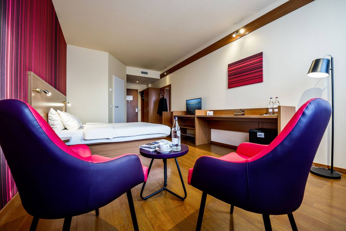 A modern hotel room with a bed, two chairs, a small table with a bottle and glasses, a desk with a computer, and a lamp.