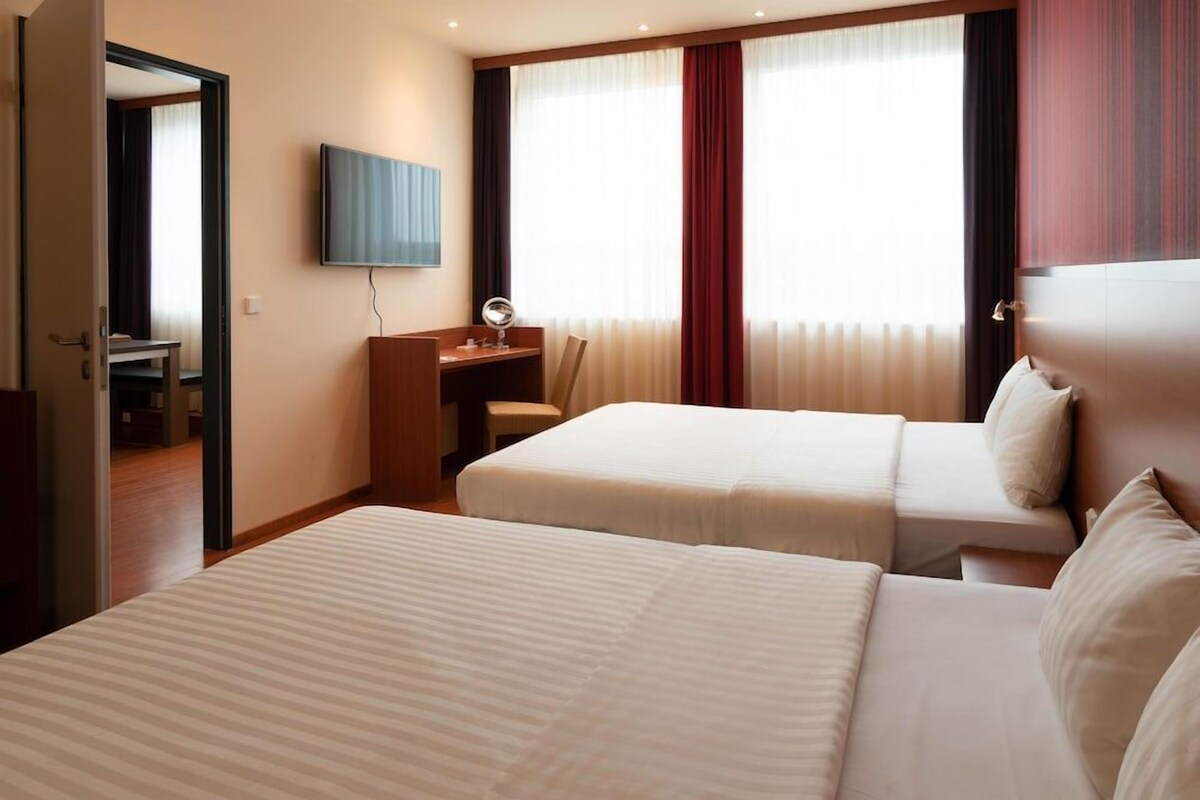 A hotel room with two beds, a desk, a chair, a television, and a window with curtains.