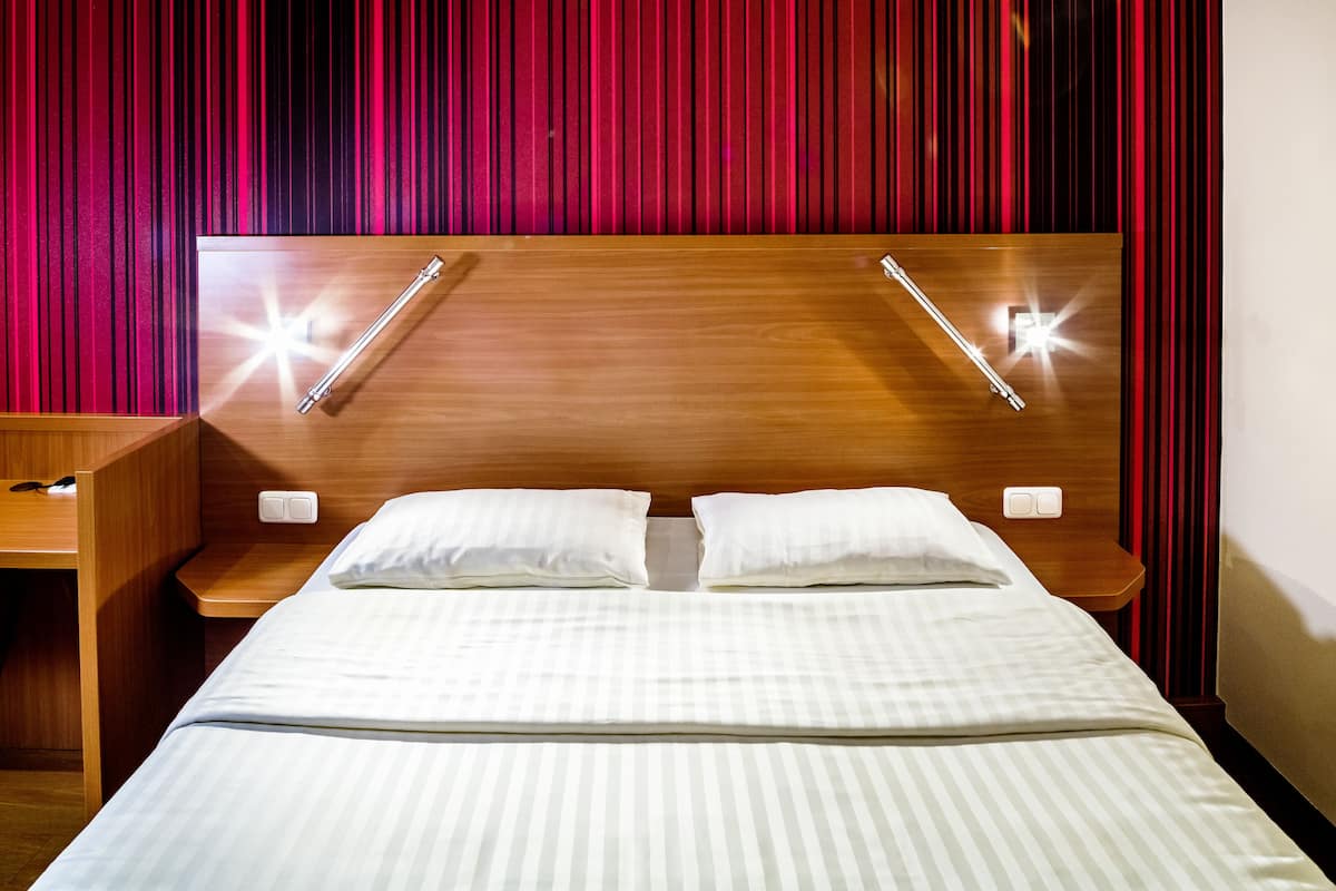 A hotel room with a wooden headboard, two bedside tables with lamps, a bed with striped bedding, and red curtains.