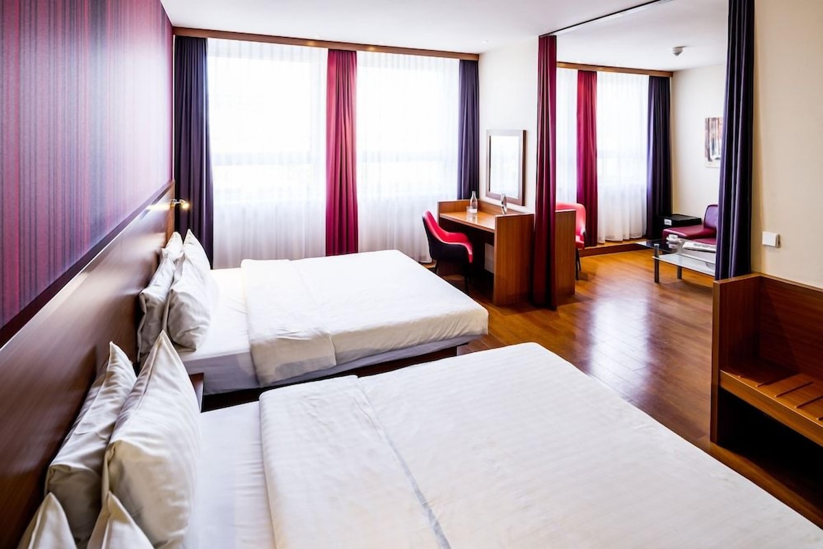 A hotel room with two beds, a desk, a chair, and a large window with curtains.