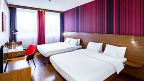 Quarto de hotel com duas camas, uma escrivaninha, uma cadeira e uma janela grande com cortinas.