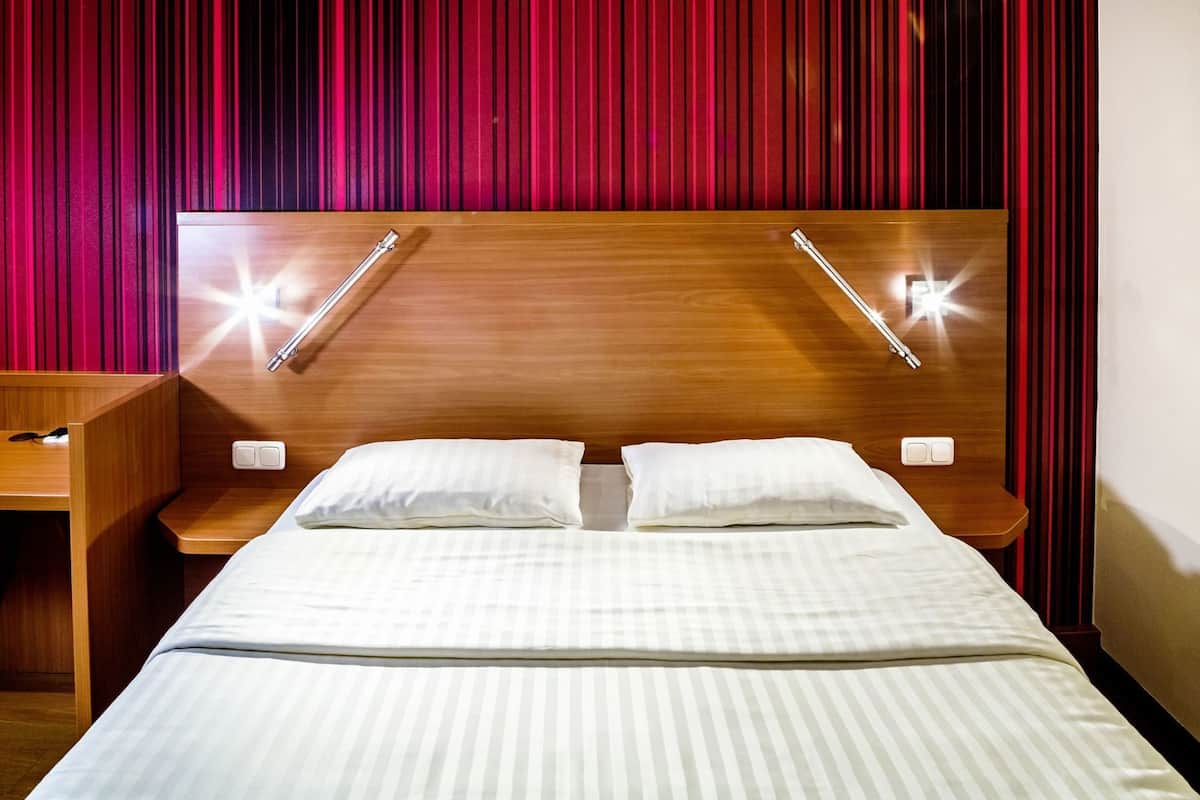 A hotel room with a wooden headboard, two bedside tables with lamps, a bed with white bedding, and a red curtain backdrop.