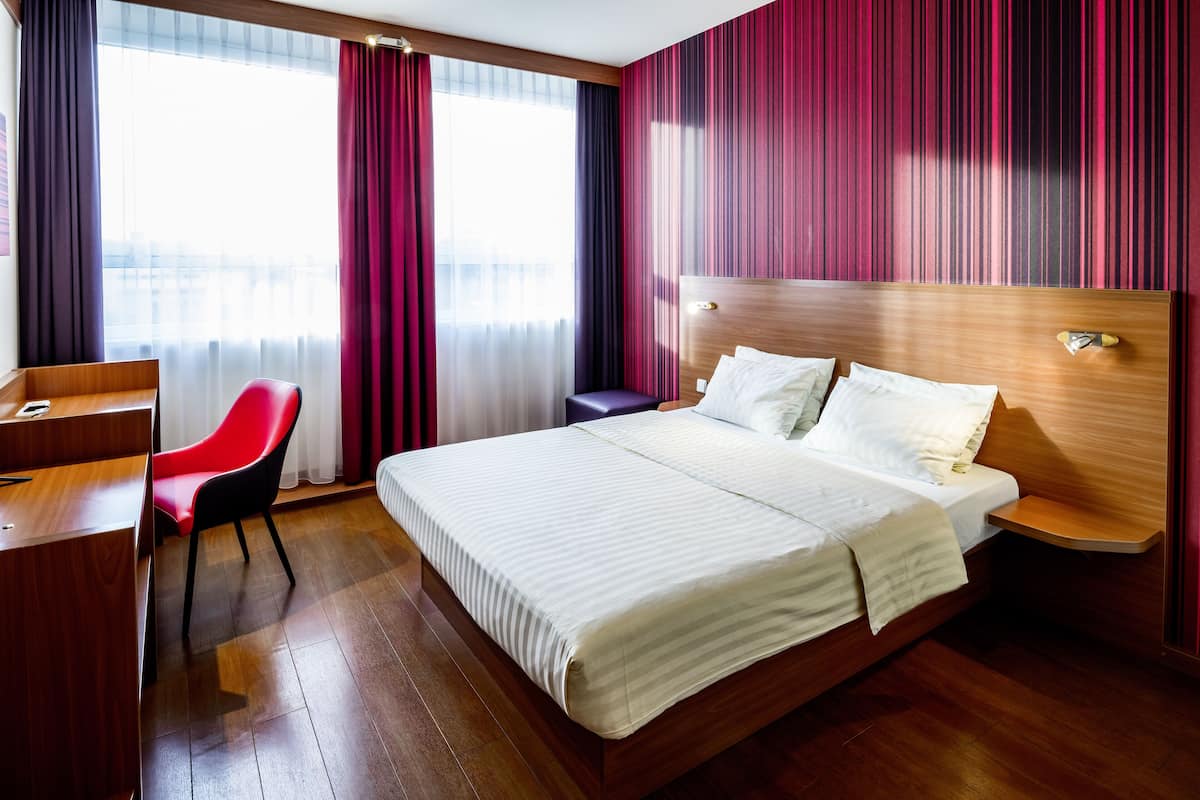A hotel room with a wooden bed, a desk, a red chair, and a view through large windows with curtains.
