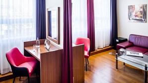 Quarto de hotel moderno com escrivaninha de madeira, duas cadeiras vermelhas, mesa de centro de vidro, sofá vermelho e um quadro na parede.