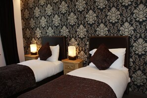 A hotel room with two beds, each with a headboard, a bedside table, and a lamp. The walls have a patterned wallpaper. - Hotel de la Gare (Monthey)