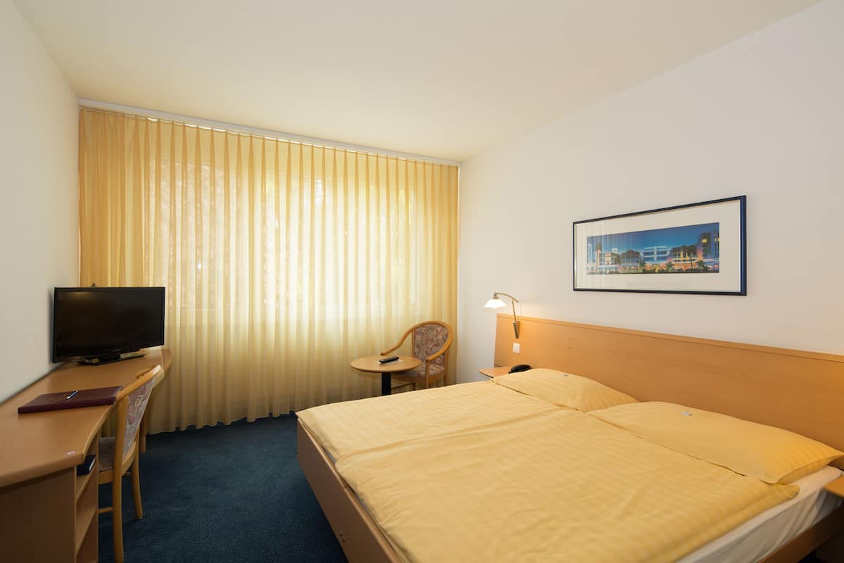 A hotel room with a bed, a desk with a television, a chair, a small table, and a wall-mounted lamp.
