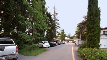 Fila de coches estacionados a lo largo de una calle residencial bordeada de árboles y un edificio blanco.