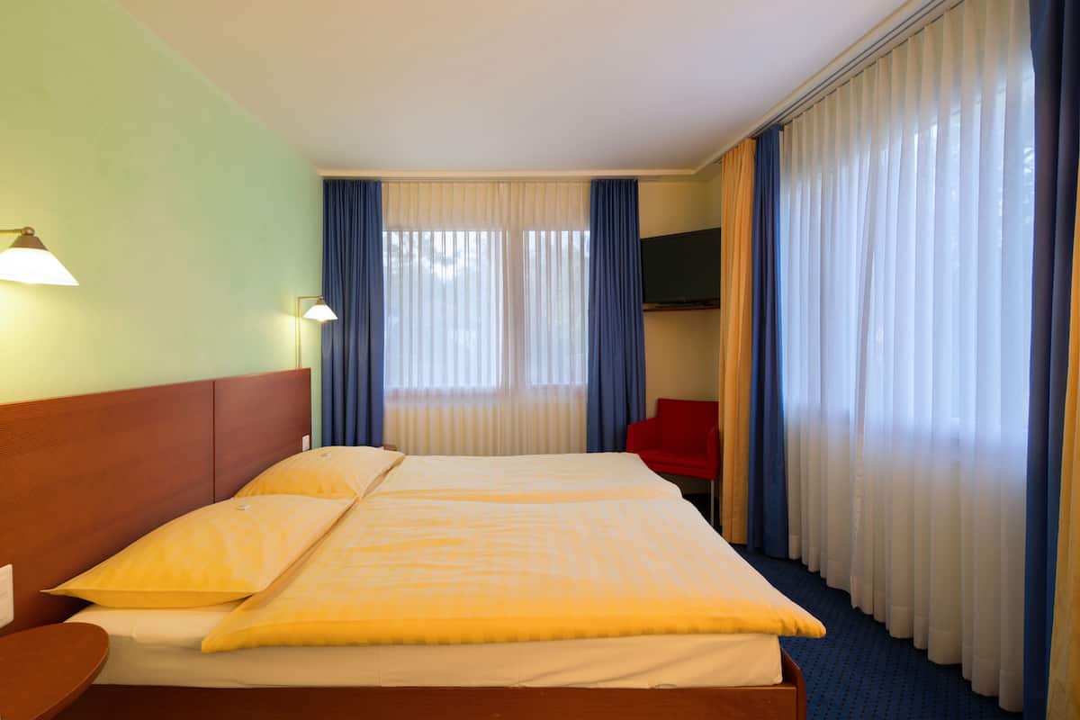 A hotel room with a double bed, a flat-screen TV, a red chair, and a large window with curtains.