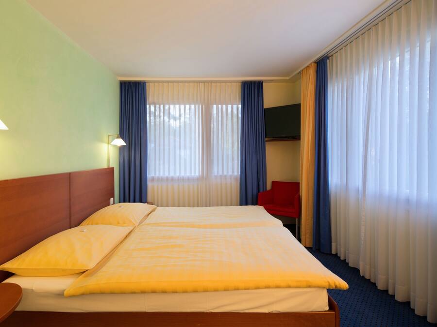 Ein Hotelzimmer mit Doppelbett, Flachbildfernseher, rotem Sessel und großem Fenster mit Vorhängen.