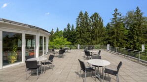 Une terrasse extérieure avec des tables et des chaises rondes, entourée de murs de verre et offrant une vue sur une forêt dense.
