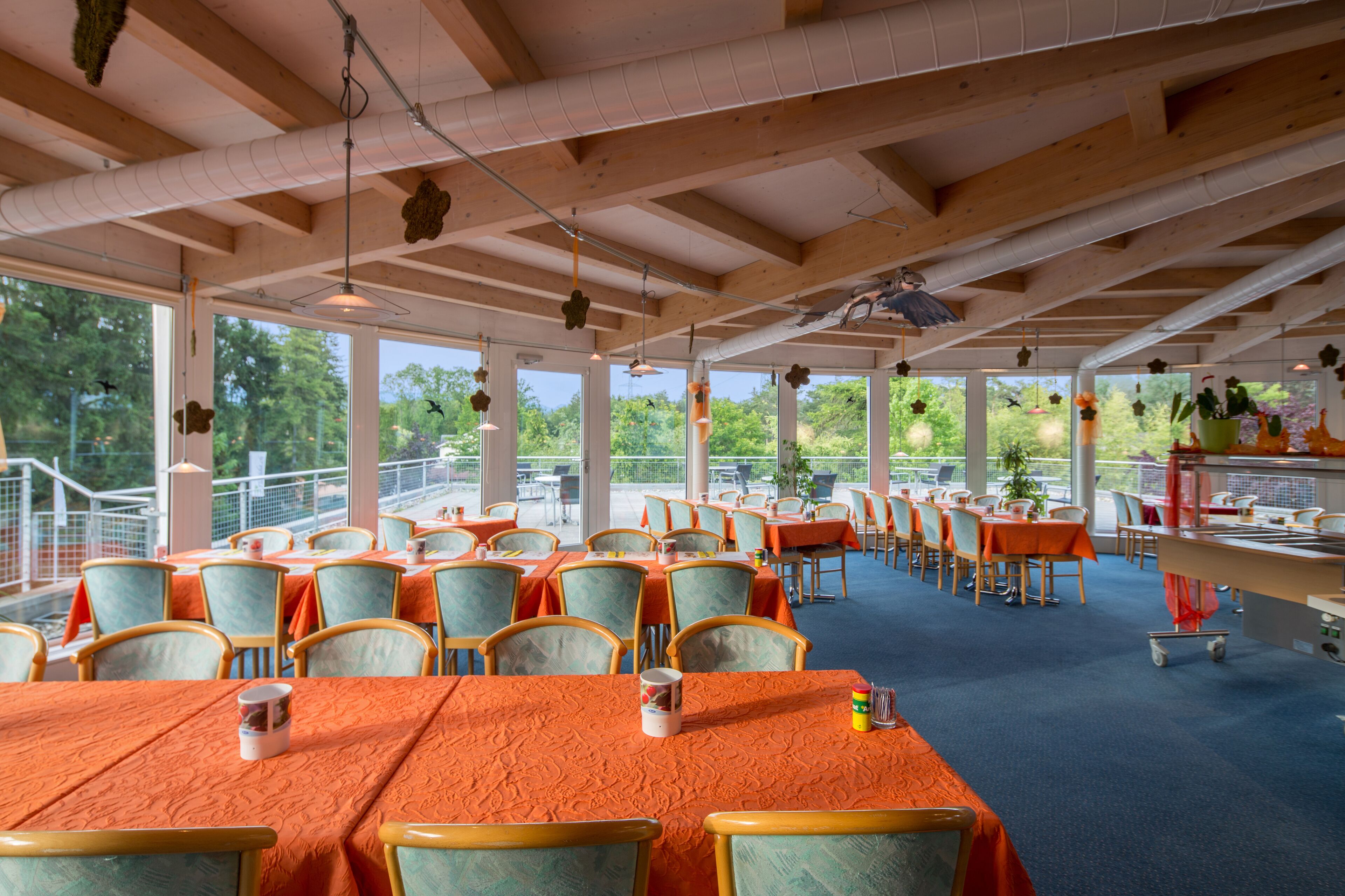 A spacious restaurant with high ceilings and large windows, featuring tables with orange tablecloths and green chairs.