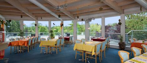 A covered outdoor dining area with multiple tables and chairs, large windows, and a view of the surrounding greenery.