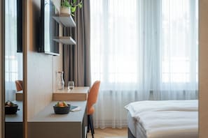 A hotel room with a bed, a desk with a chair, a plant, a vase, and a window with sheer curtains. - Tailormade Hotel METROPOL St.Gallen (St. Gallen)