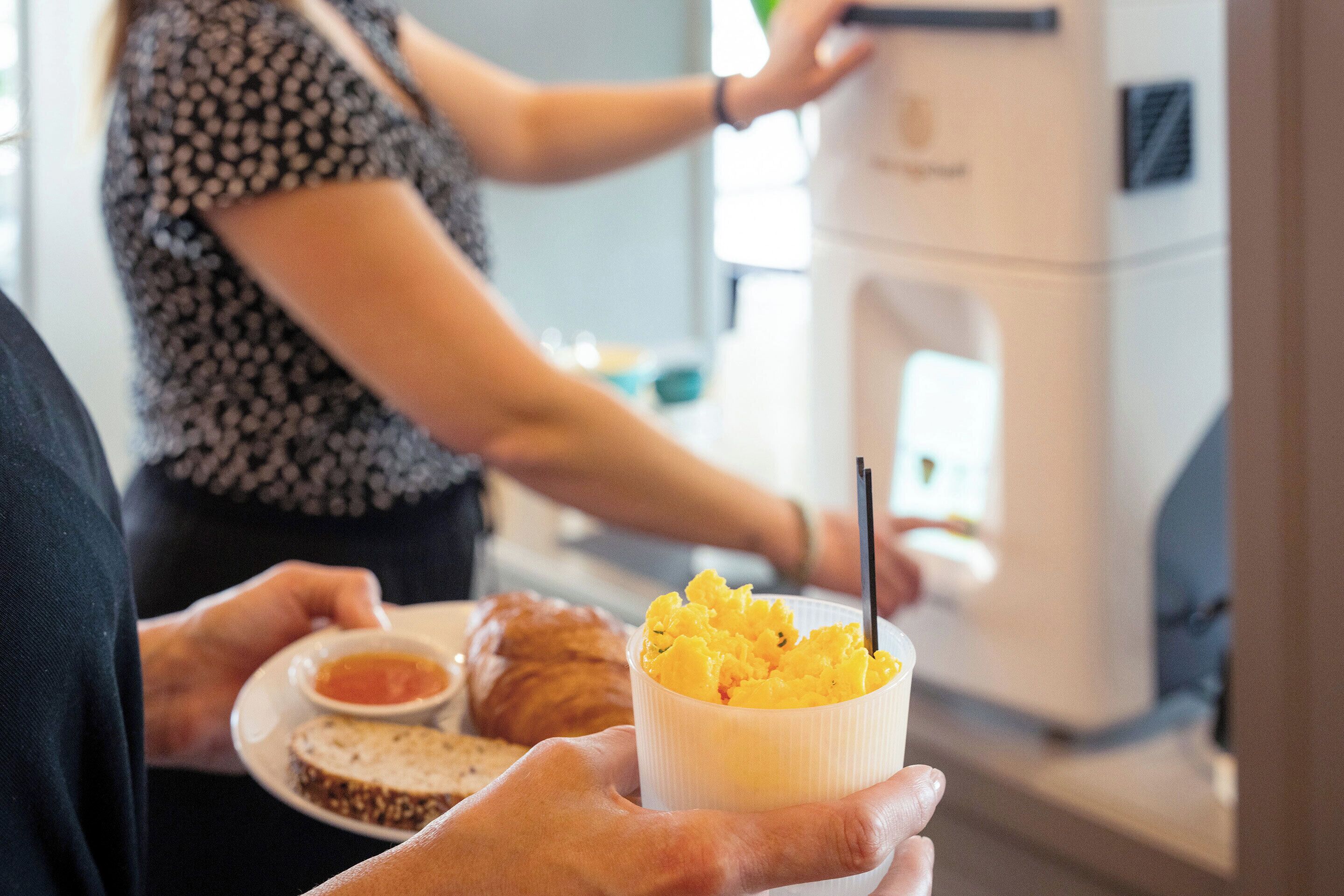 A person holding a cup of food with a spoon, a plate with a croissant and a small bowl of sauce, and a coffee machine in the background.