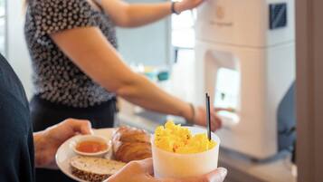 A person holding a cup of food with a spoon, a plate with a croissant and a small bowl of sauce, and a coffee machine in the background.