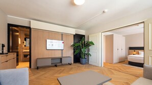 A modern hotel room with a wooden floor, a grey sofa, a small table, a large potted plant, and a bed with a headboard. - Tailormade Hotel METROPOL St.Gallen (St. Gallen)