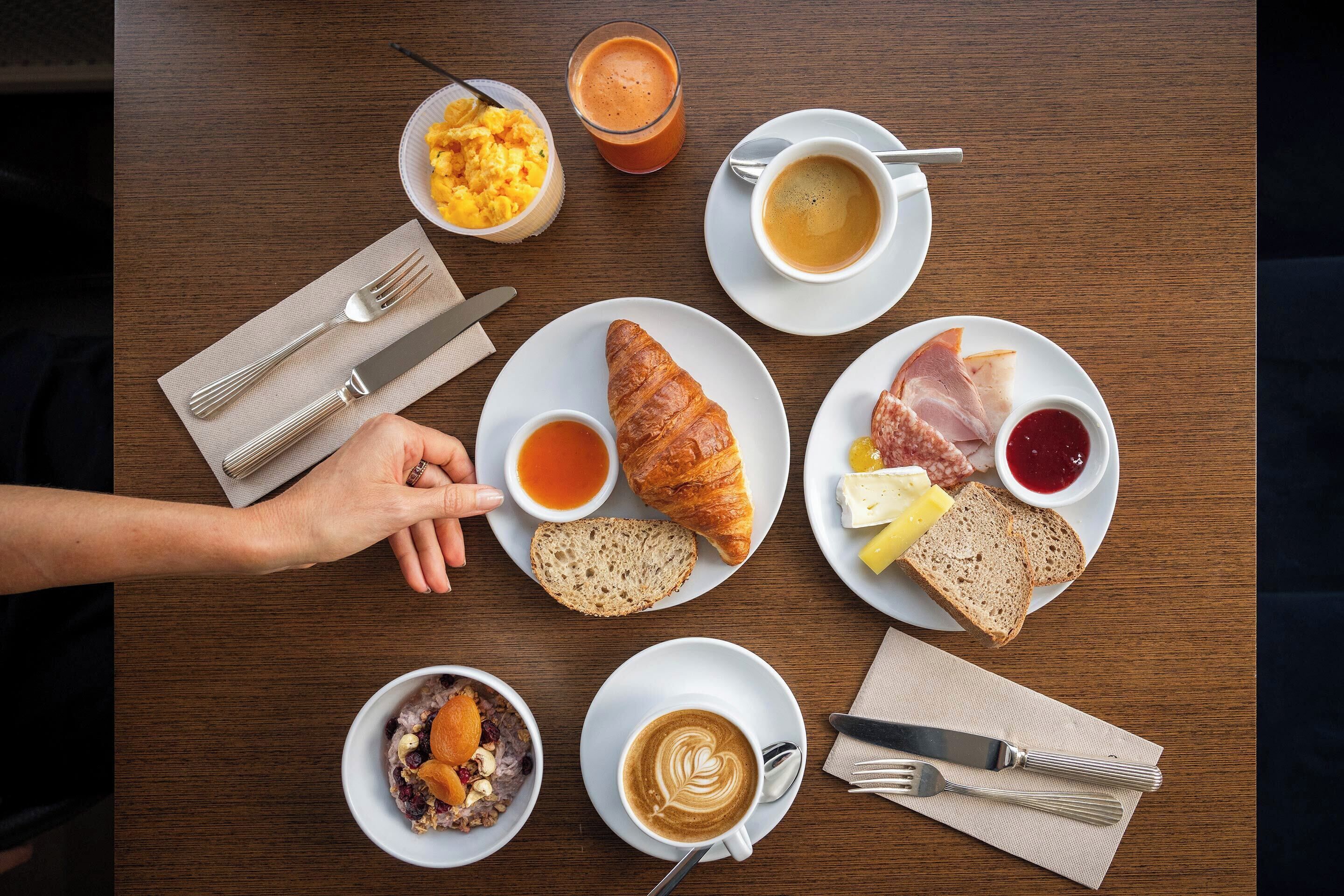 Une table dressée pour le petit-déjeuner avec des croissants, du pain, du fromage, du jambon et diverses confitures sur des assiettes, ainsi que des tasses de café et de jus de fruits.