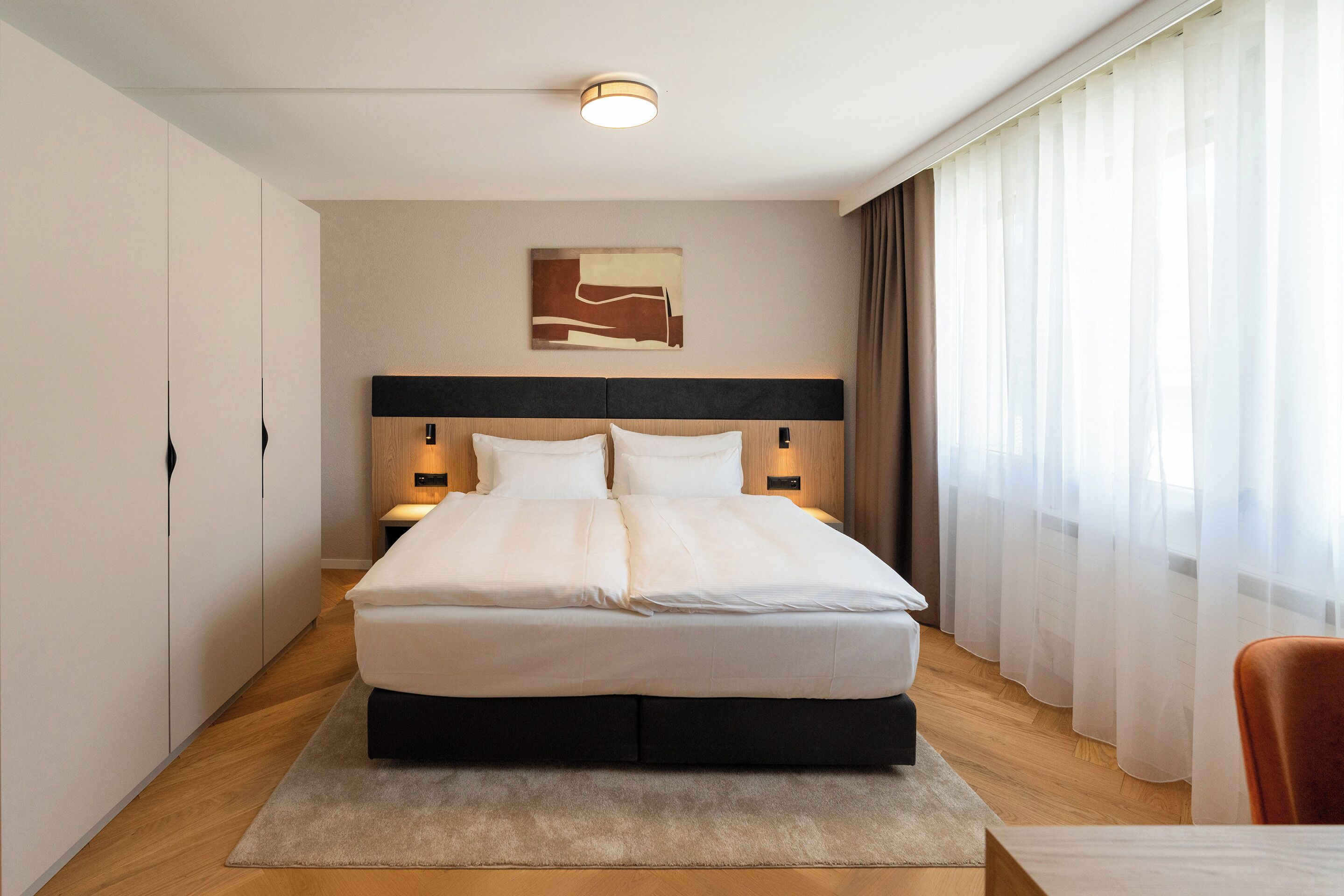 Una camera d'hotel moderna con un letto grande, lampade sui comodini, una testiera e vista sull'esterno attraverso tende leggere.