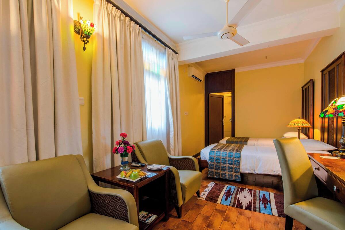 Ein Hotelzimmer mit einem Bett, zwei Stühlen, einem Tisch mit Blumenvase, einer Lampe und einem Deckenventilator.