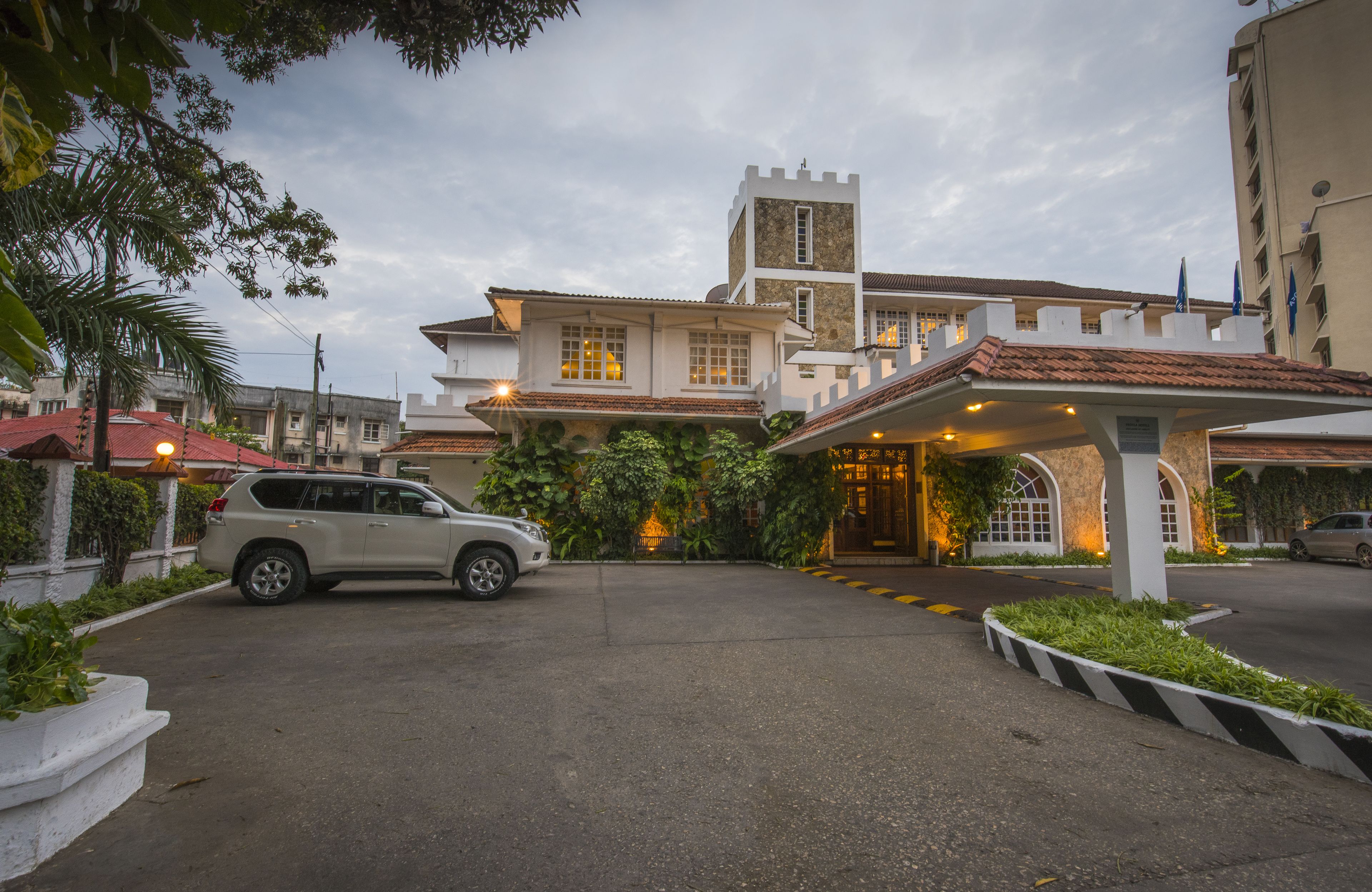 A hotel entrance with a car parked in front, a covered area with plants, and a building with a tower.