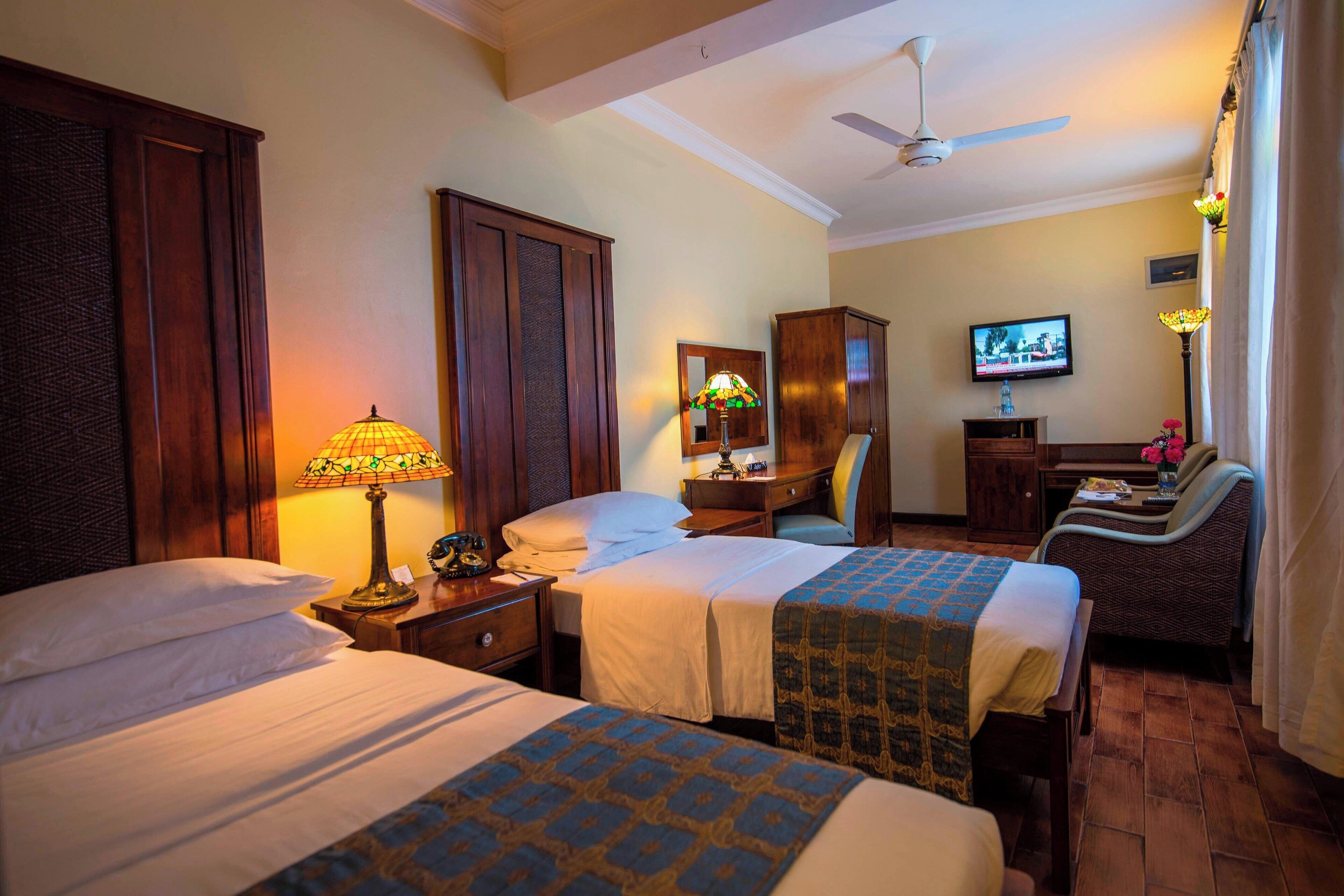 A hotel room with two beds, a television, a desk with a chair, and a ceiling fan.