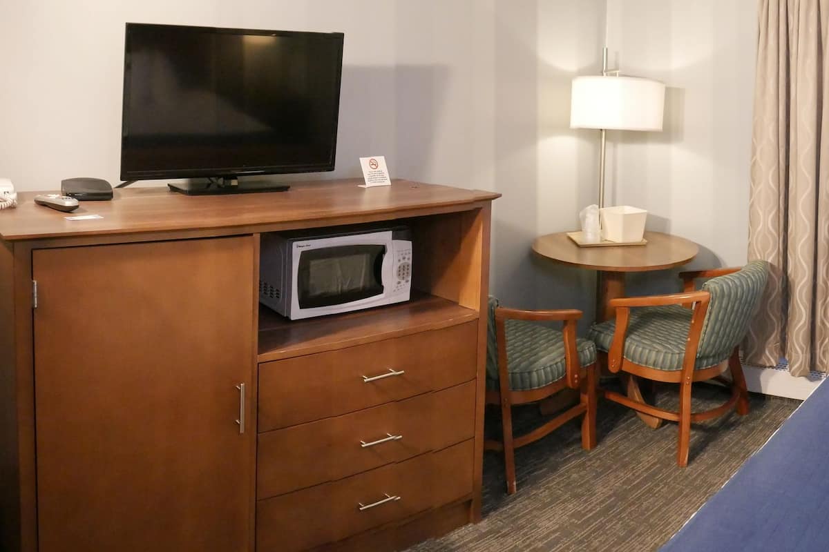 A hotel room with a bed, a desk with a television, a microwave, a chair, a small table with a lamp, and a window with curtains.
