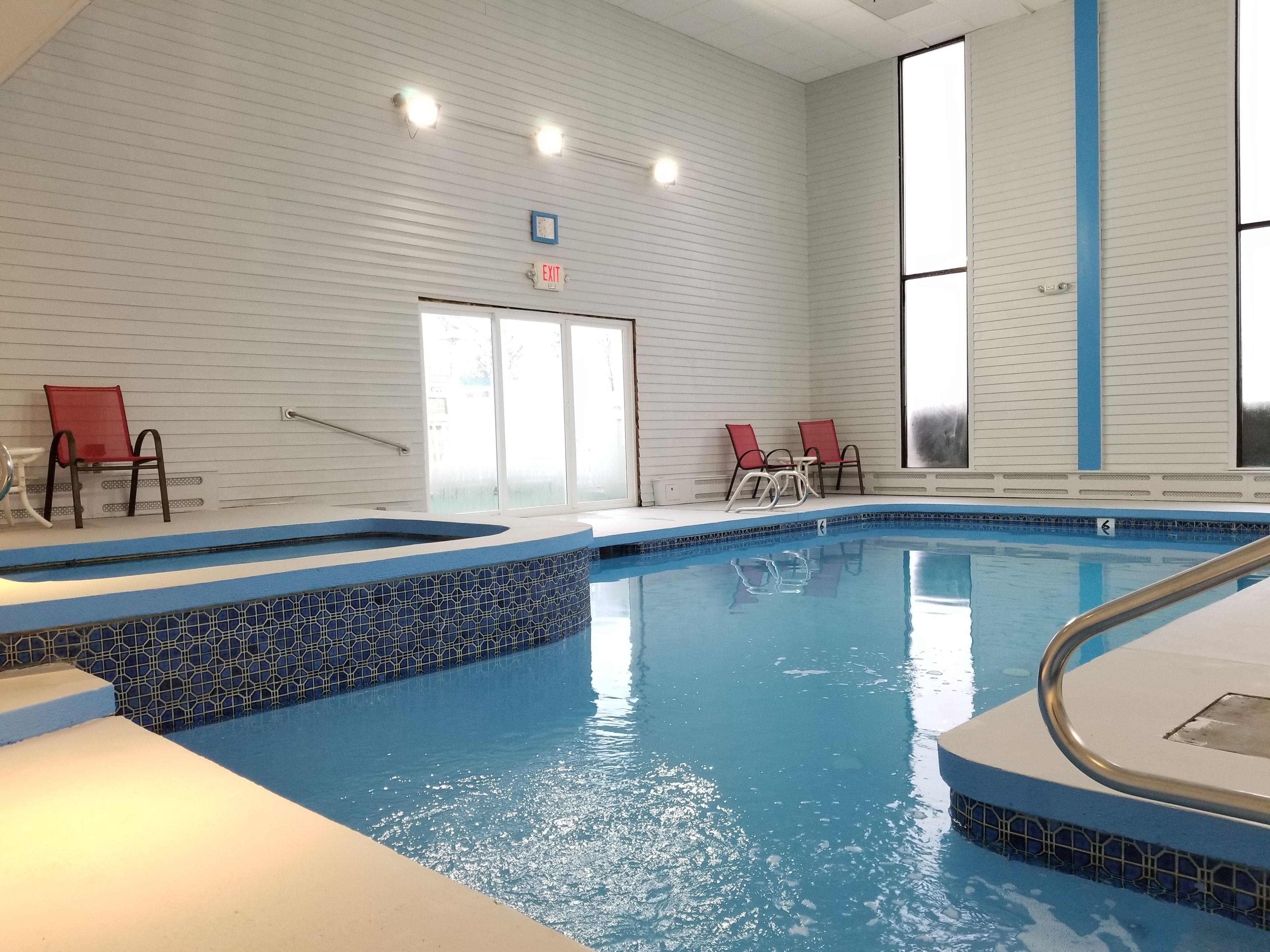 An indoor swimming pool with a tiled edge, two red chairs, and an exit sign on the wall.