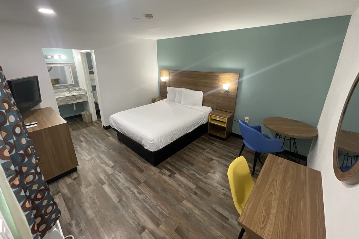 A hotel room with a bed, bedside tables, a desk with a chair, a television, and a bathroom visible through an open door.