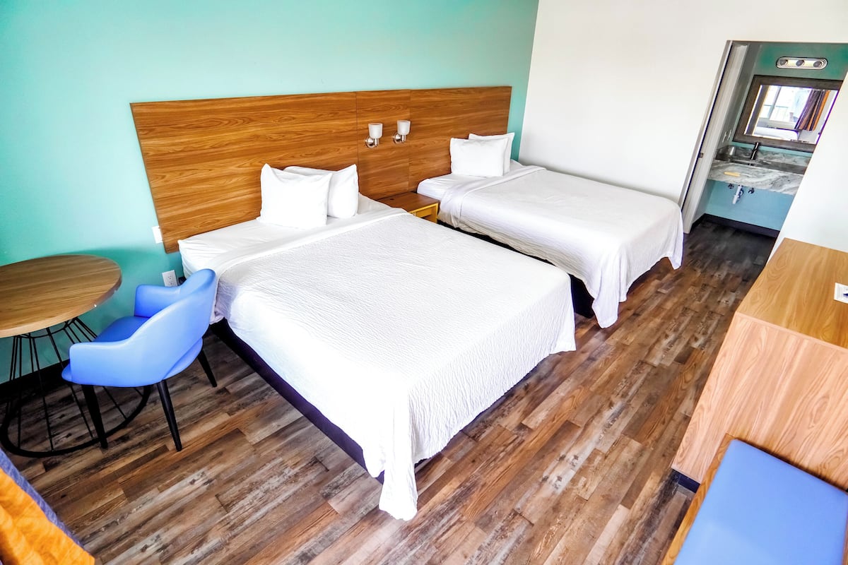 A hotel room with two beds, a wooden headboard, a small table with a chair, and a view of the bathroom.