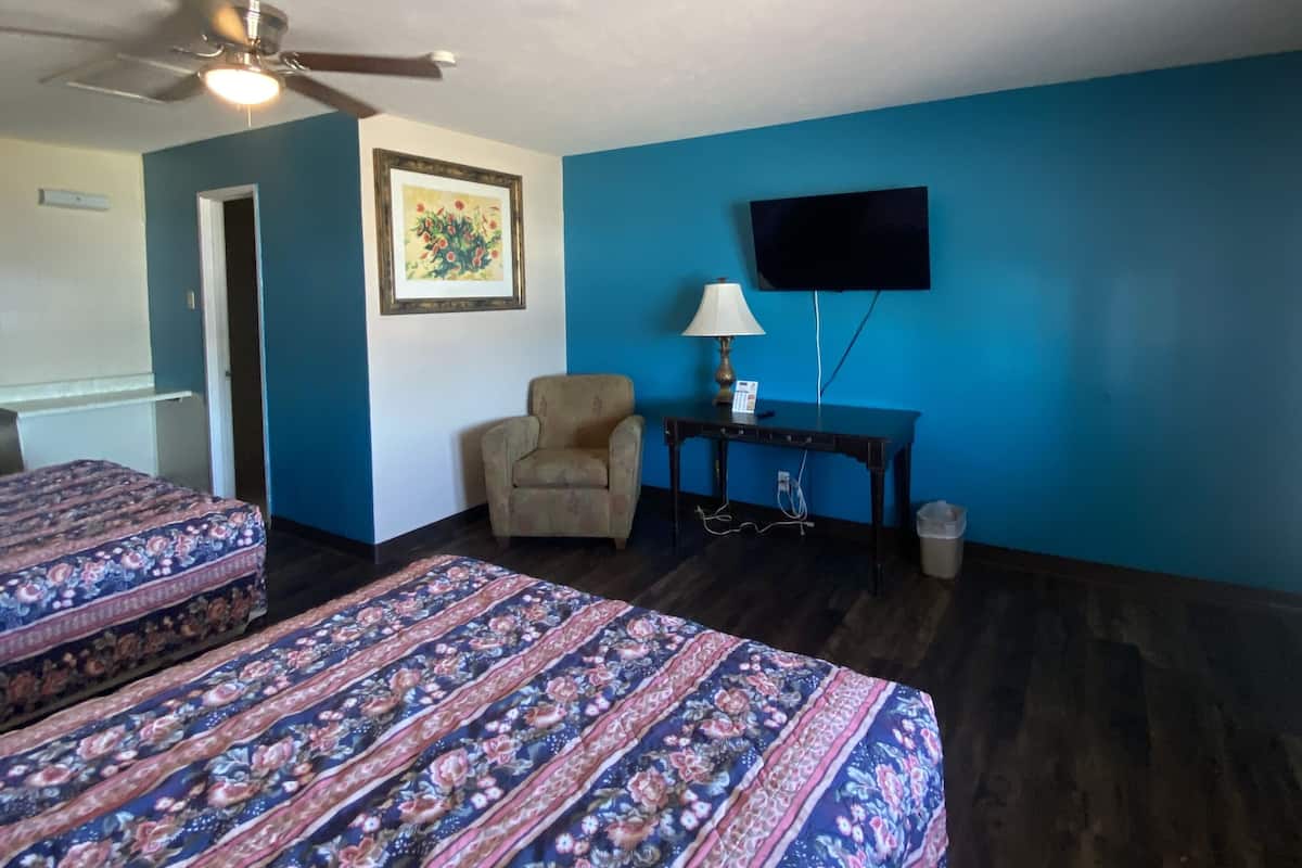 A room with two beds, a ceiling fan, a TV, a chair, and a desk with a lamp.