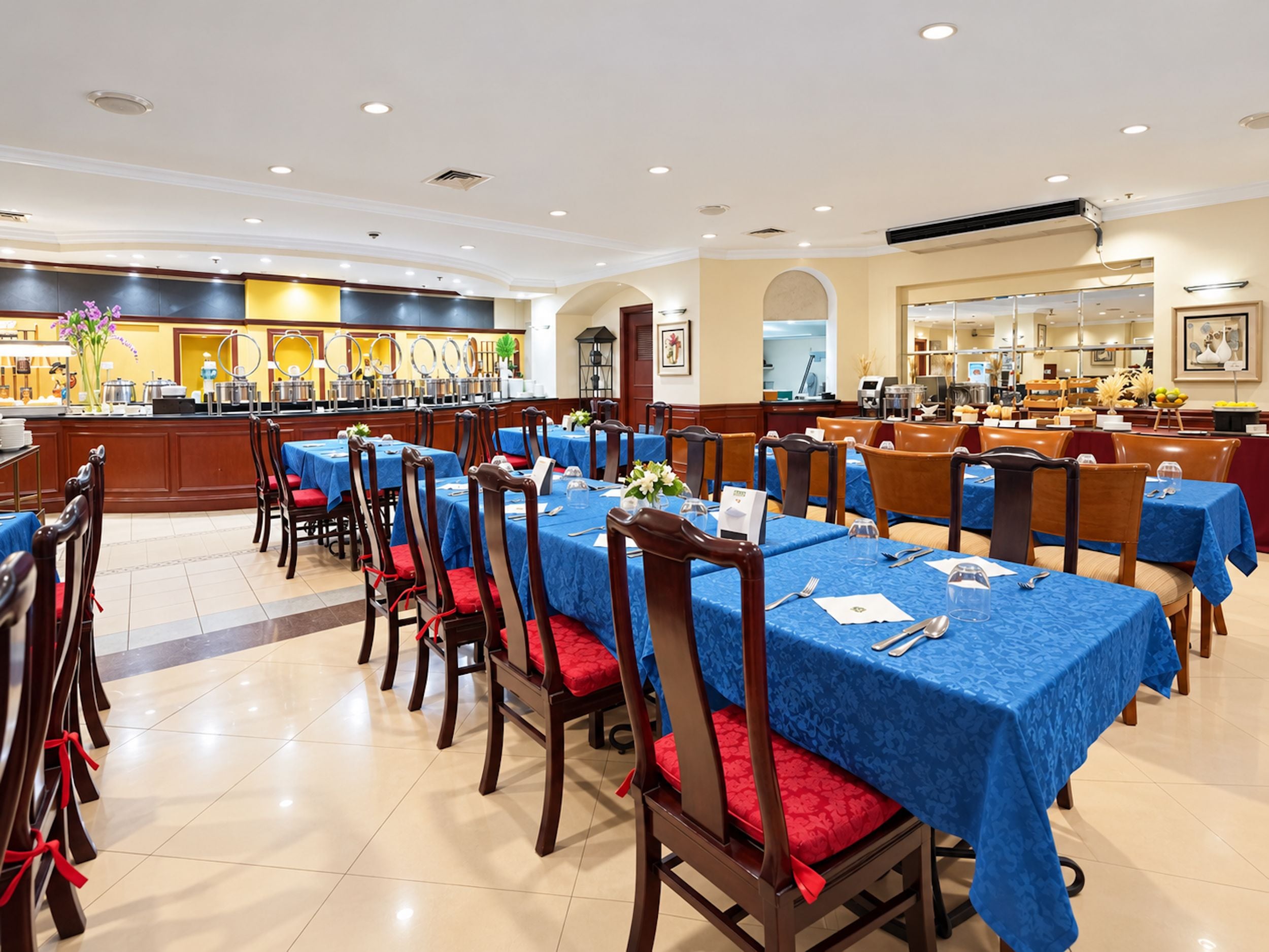 A hotel dining area with multiple tables and chairs, a buffet counter, and a kitchen in the background.