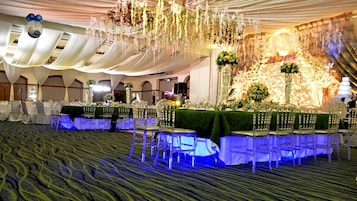 Un salón de banquetes con mesas redondas cubiertas con manteles blancos, sillas con fundas verdes y un escenario con adornos florales.