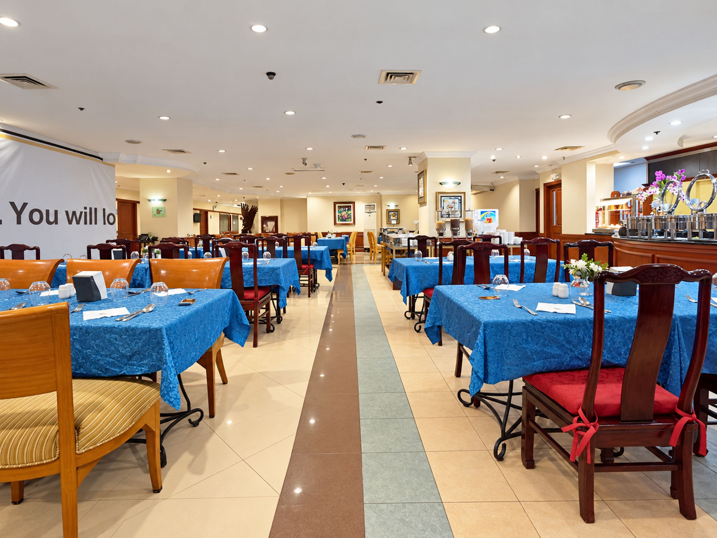 A hotel dining area with multiple tables and chairs, a reception desk, and a banner.