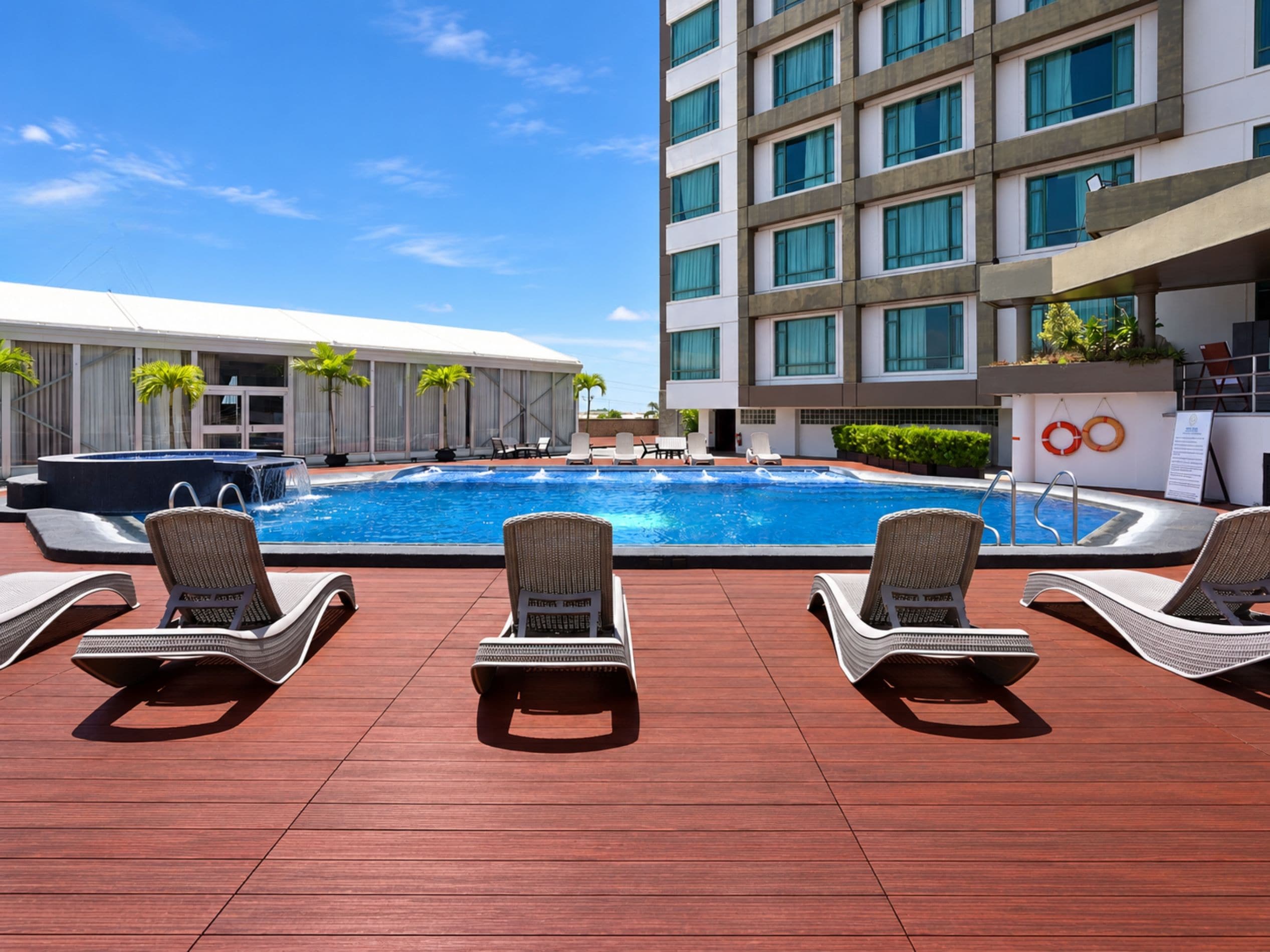 Zona de piscina de hotel con sillones reclinables y un edificio al fondo.