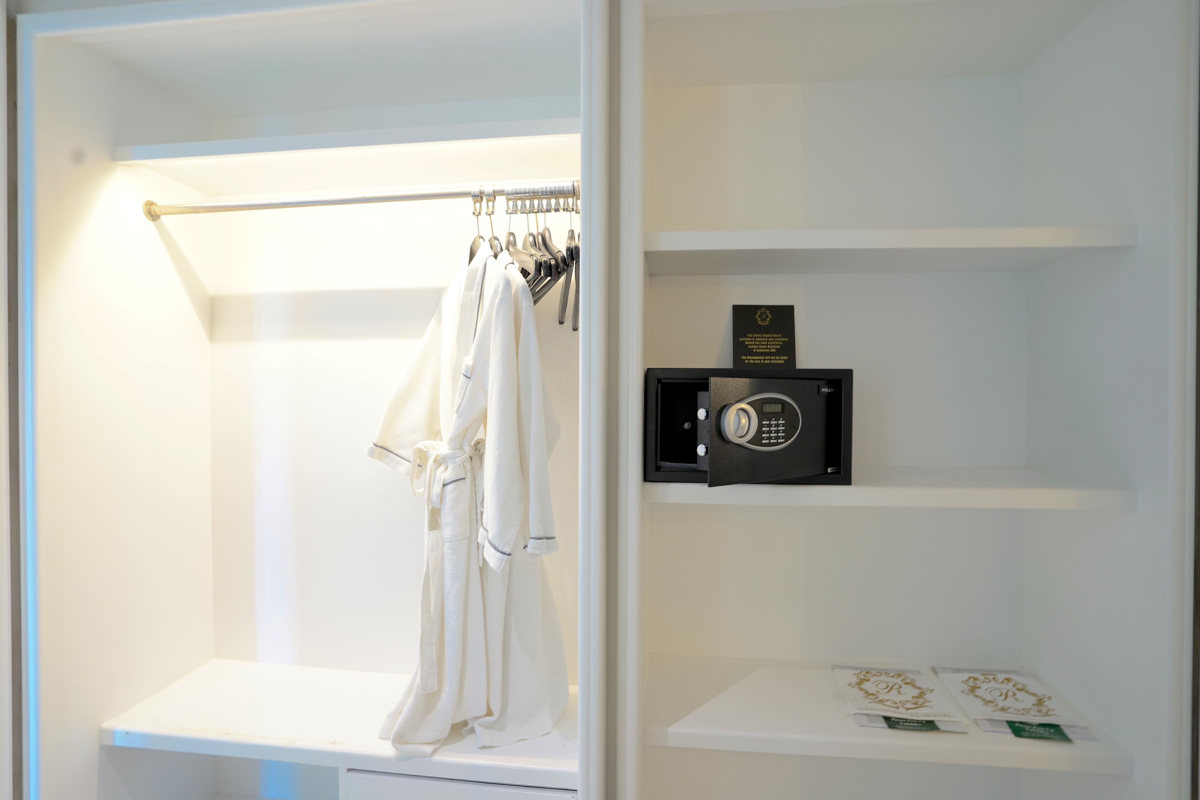 A white closet with hanging robes, a safe, and a shelf with items.