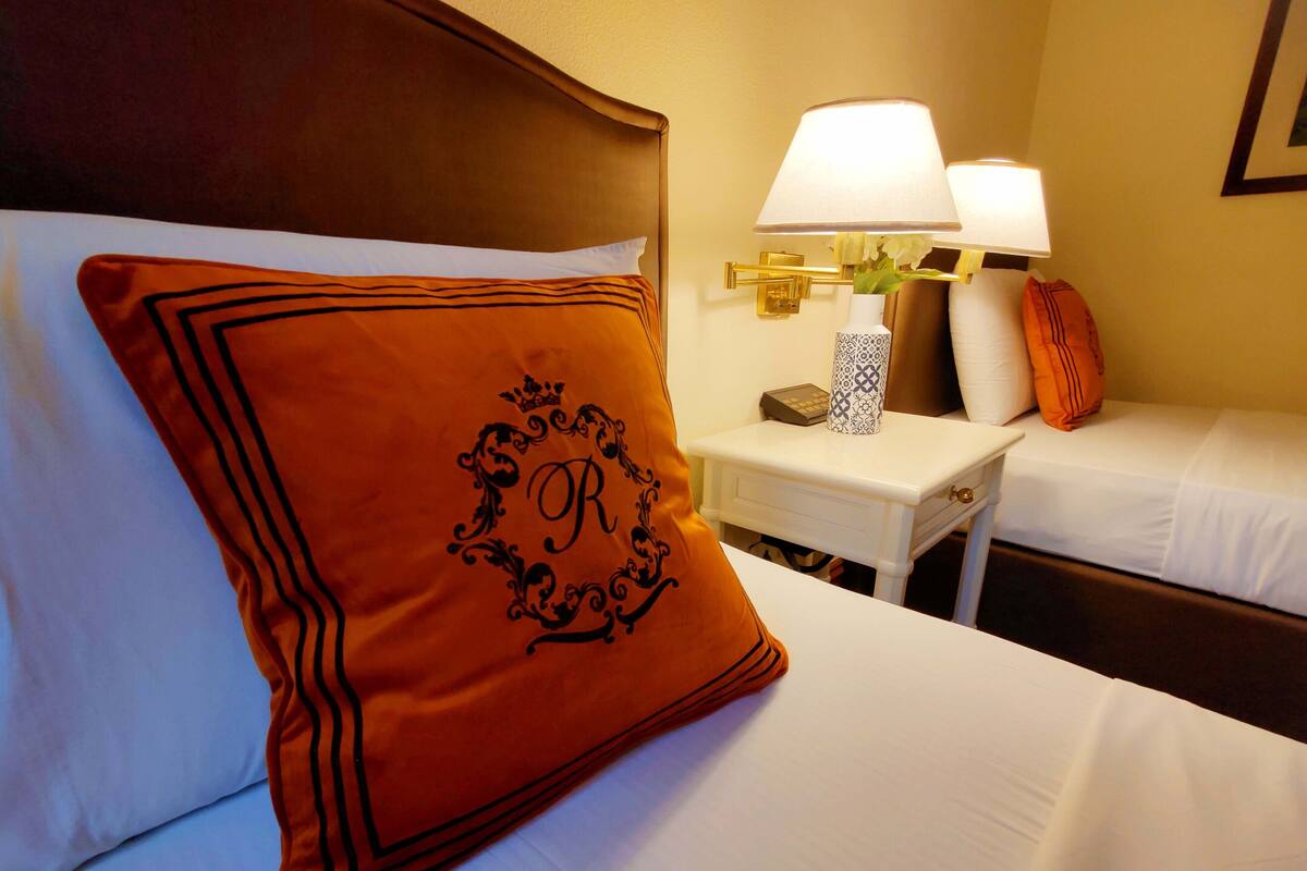 Une chambre d’hôtel avec deux lits, une table de chevet avec des lampes et un coussin décoratif orné d’un monogramme.