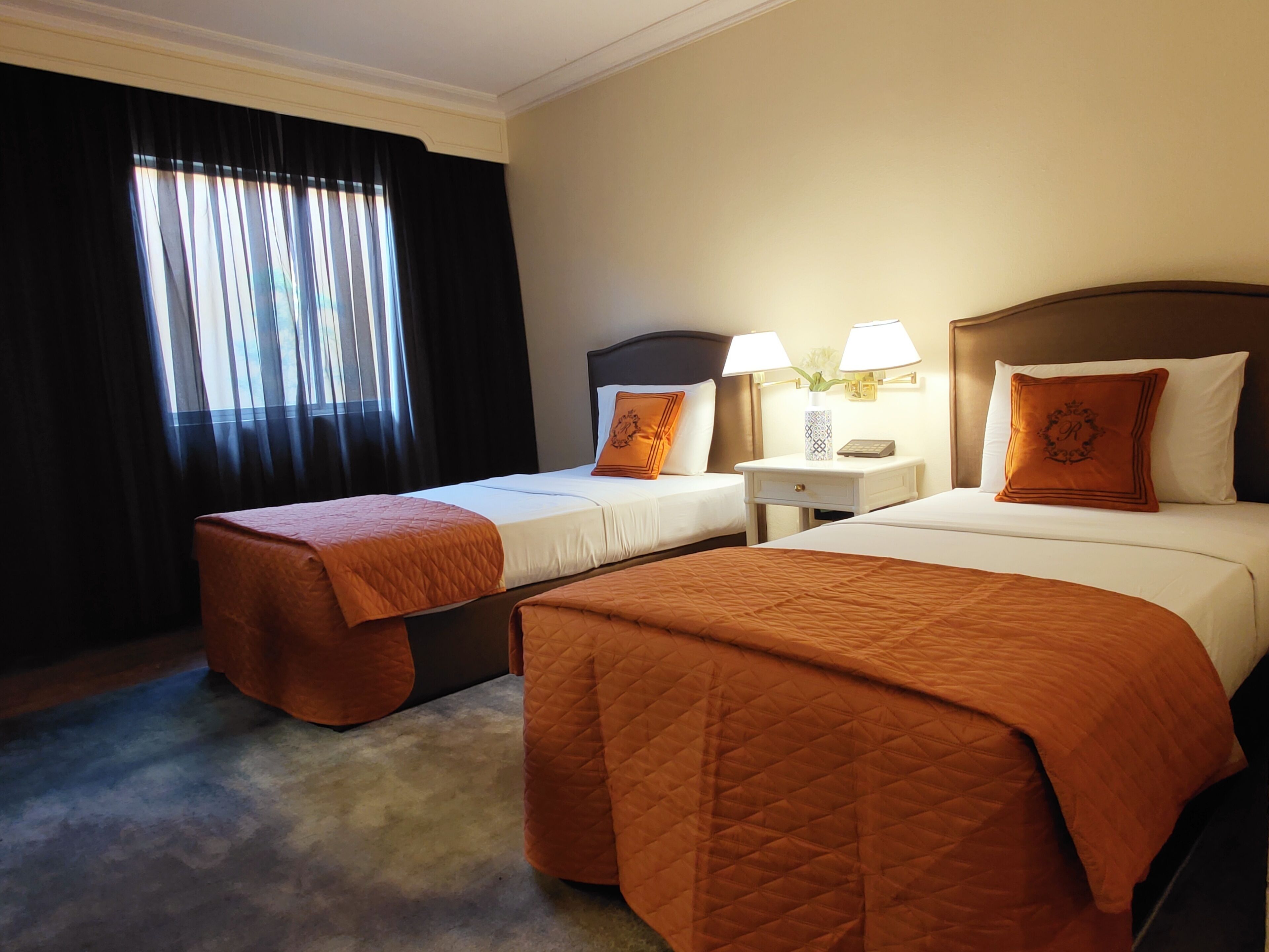 Habitación de hotel con dos camas, cada una con una colcha naranja acolchada, una mesita de noche con dos lámparas y una ventana con cortinas negras.