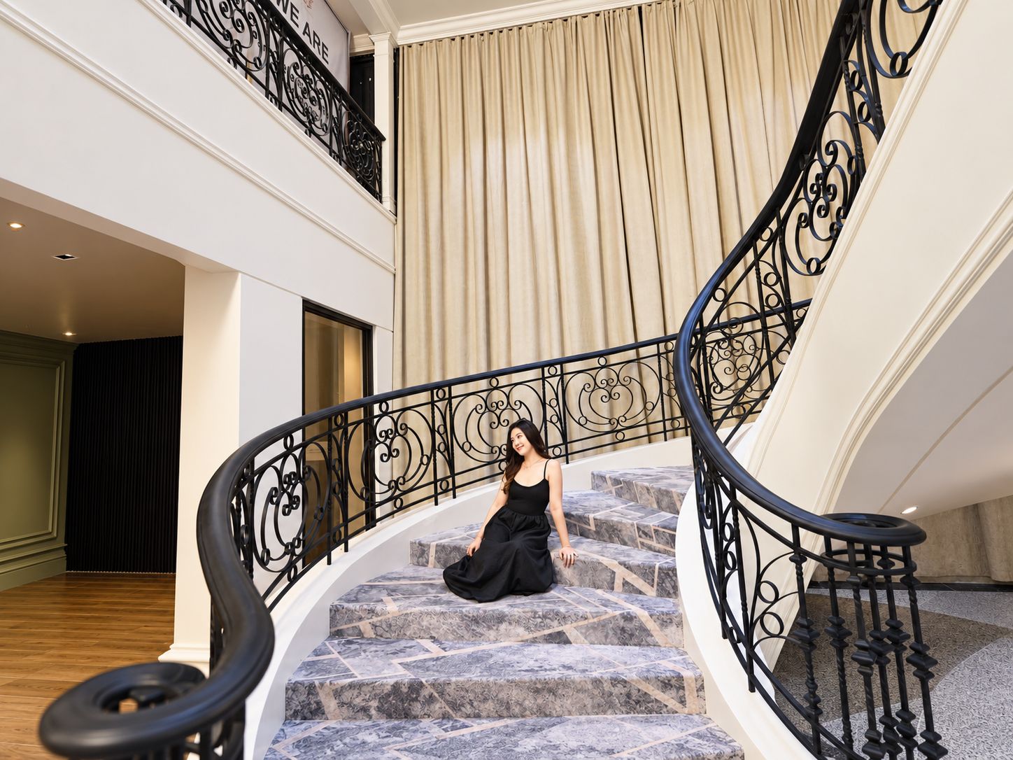 A woman sitting on a curved staircase with a black railing, leading to an upper floor.