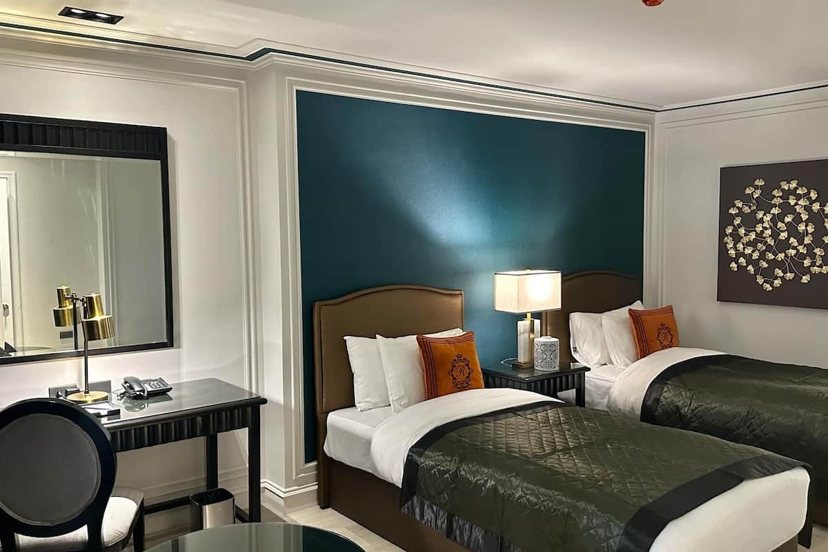 Une chambre d’hôtel avec deux lits, un bureau, une chaise, un miroir et une table de chevet avec une lampe.