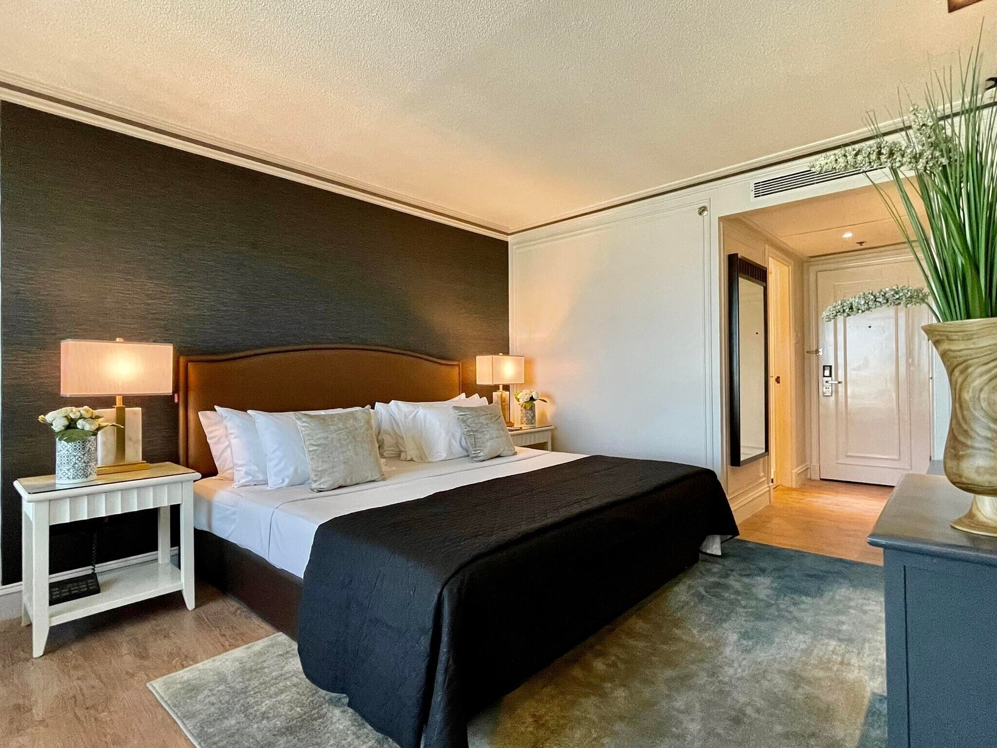 Habitación de hotel con una cama grande, dos mesitas de noche, una mesilla auxiliar, una planta en maceta y una puerta que da a otra habitación.