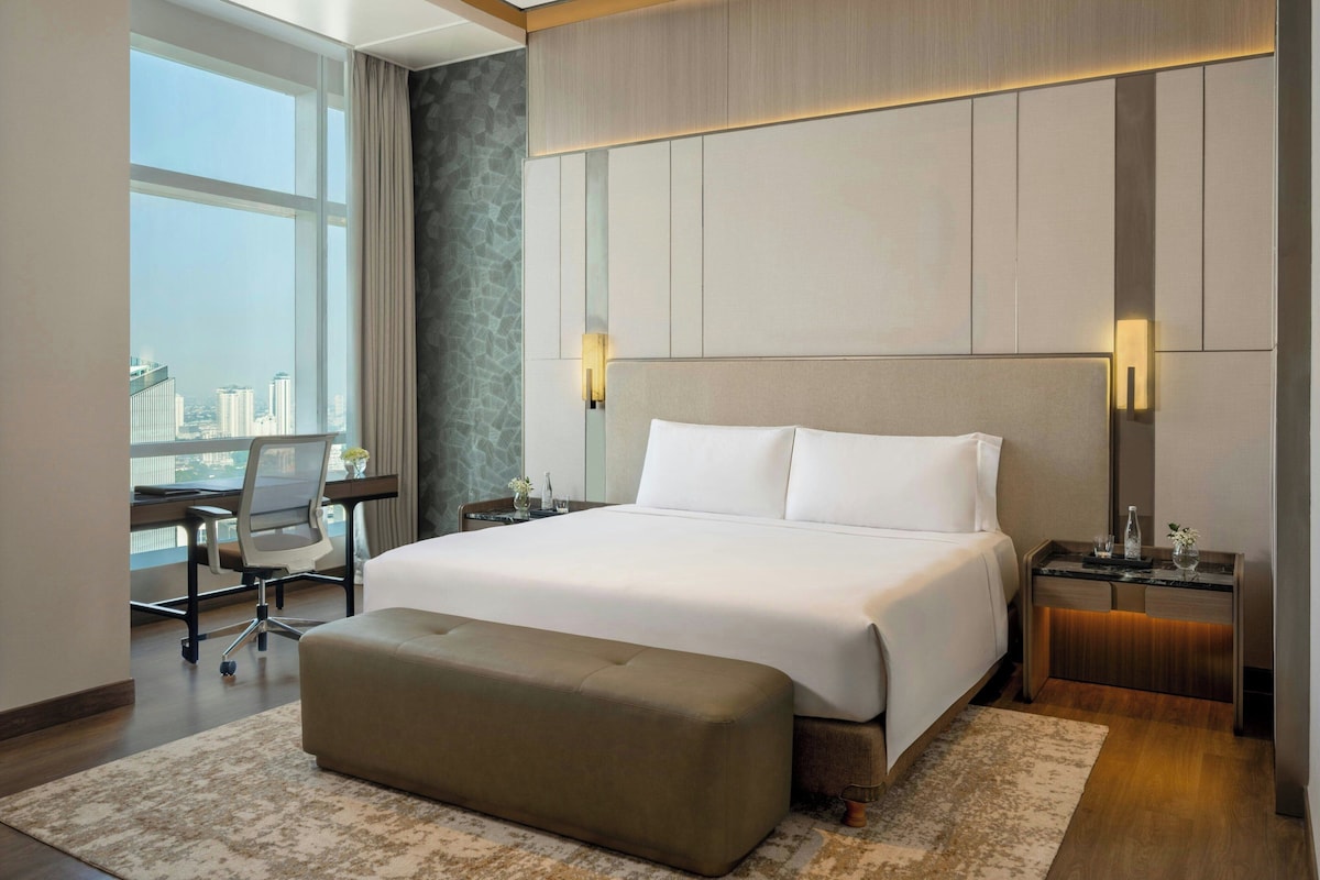 A hotel room with a large bed, a desk, a chair, and a view of the city.
