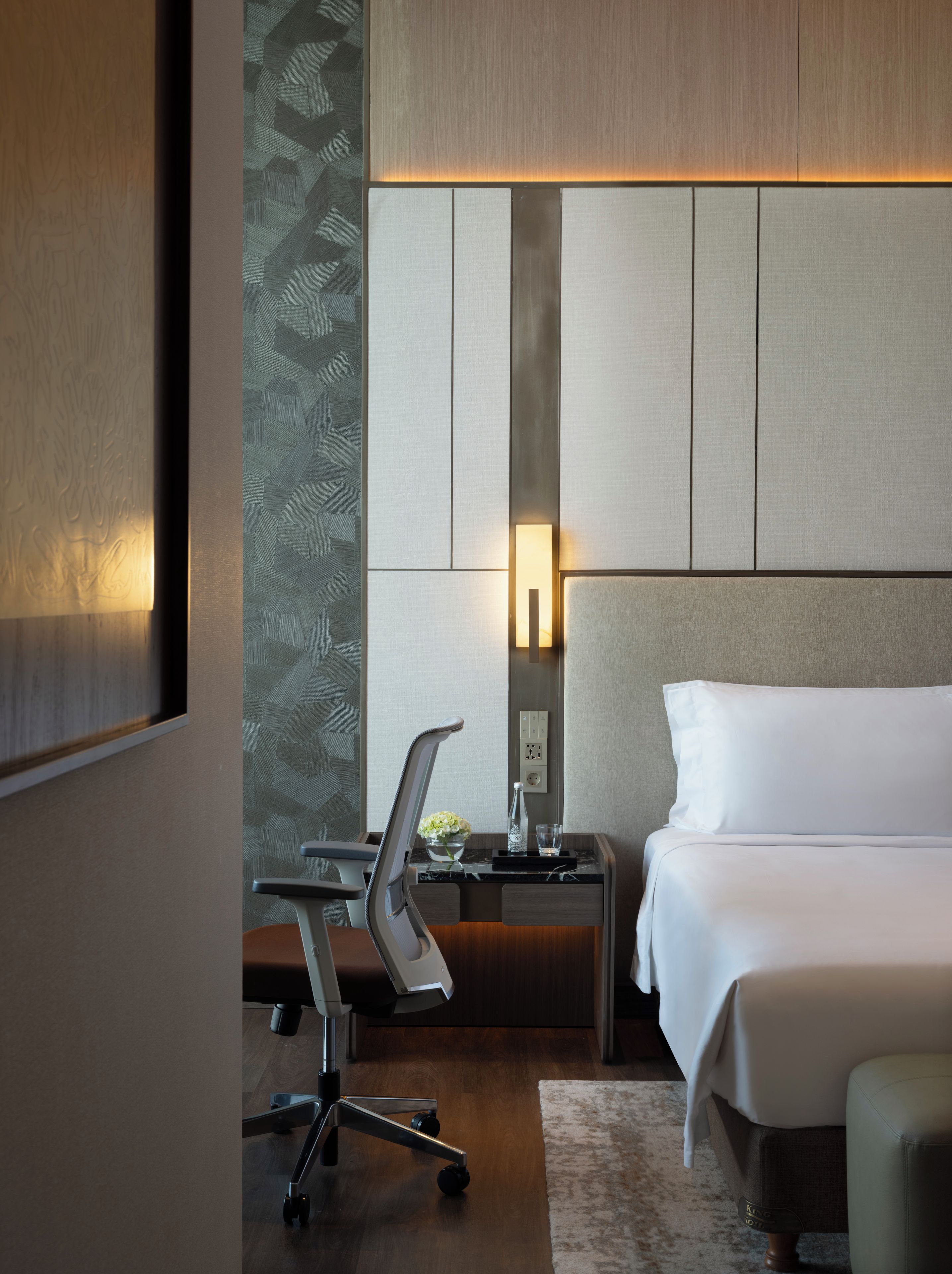 A hotel room with a bed, a desk, a chair, a nightstand, and a wall-mounted lamp.