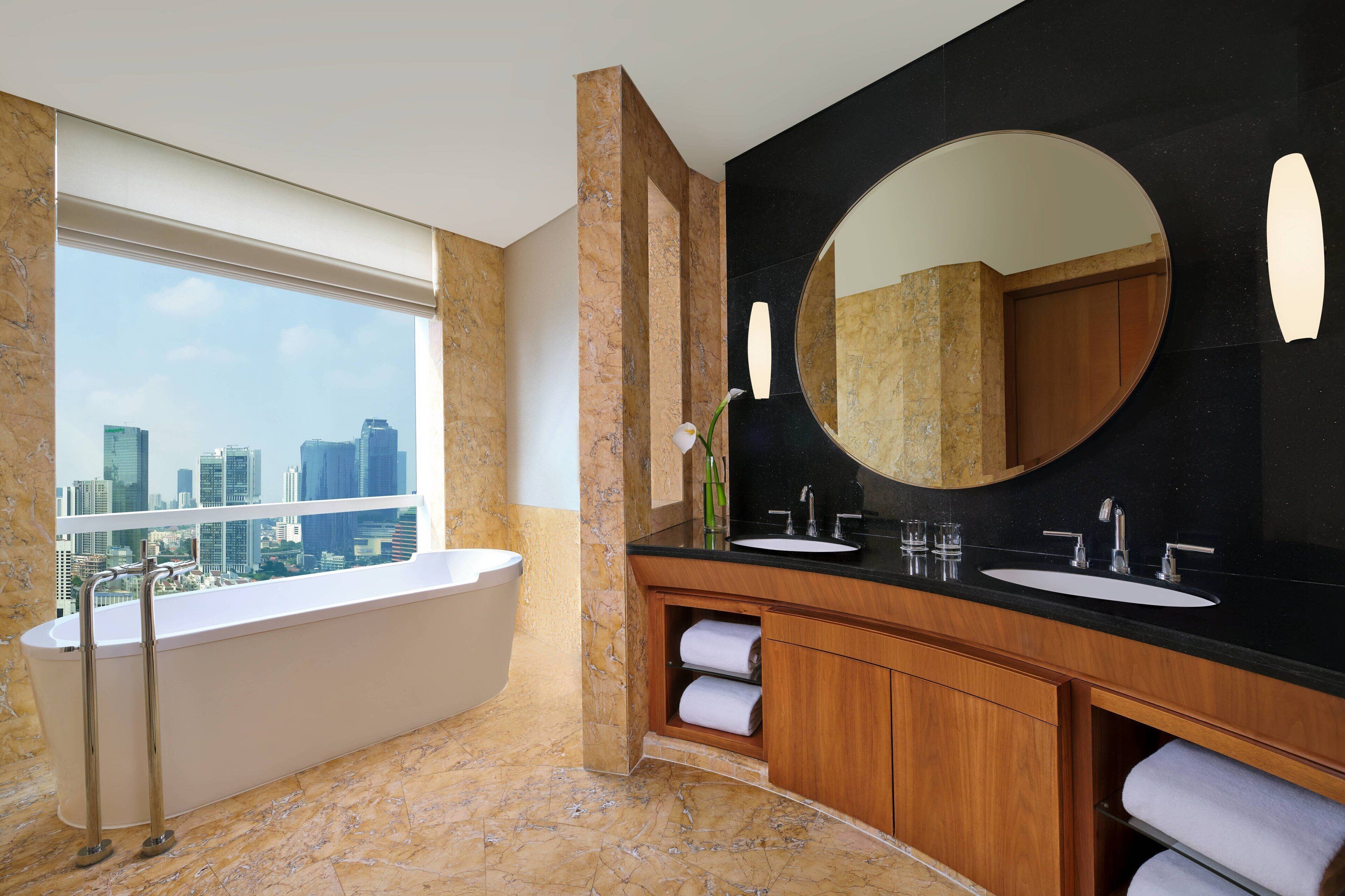 Un baño moderno con bañera, dos lavamanos, un espejo grande y vistas a la ciudad a través de un ventanal.