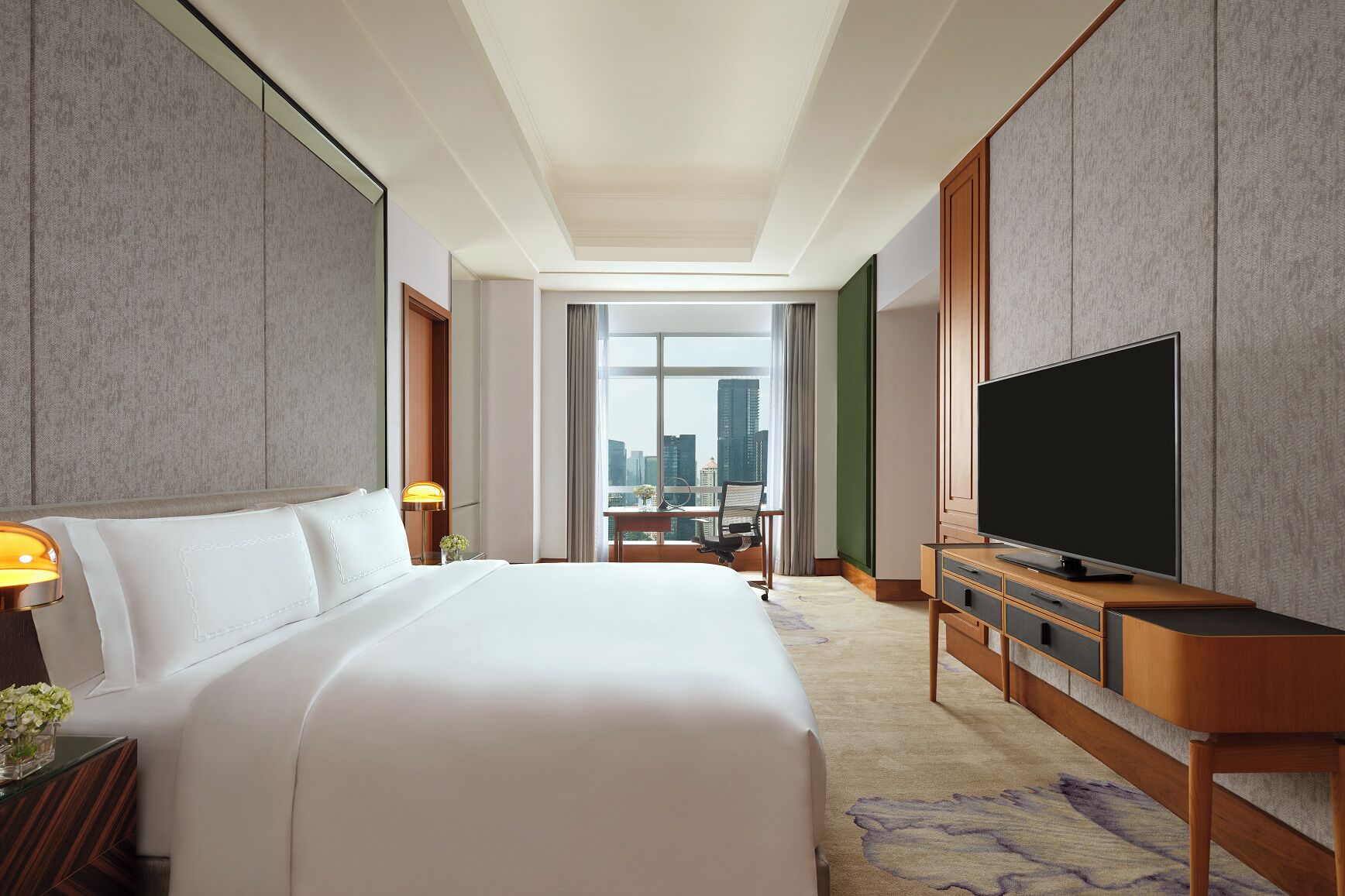 A hotel room with a large bed, a flat-screen TV, a desk with a chair, and a view of the city through a large window.