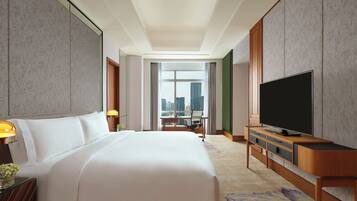 A hotel room with a large bed, a flat-screen TV, a desk with a chair, and a view of the city through a large window.
